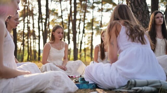 Group of young women in white dresses meditating together on grass under trees in a serene outdoor setting during daylight with soft sunlight filtering through branches