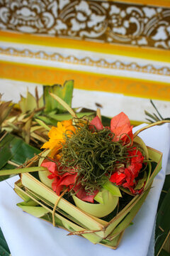 Close-up of a palm leaf offering with red and orange flowers and green fern beside a white cloth with gold patterns, Ubud, Indonesia