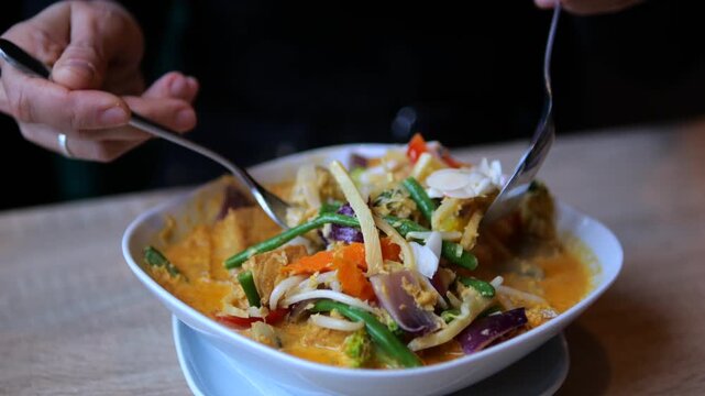 Hands stir vegetable curry with two spoons in white bowl. Colorful vegetables move through rich orange sauce, presenting vegan cuisine as diners prepare fresh meal at wooden table.