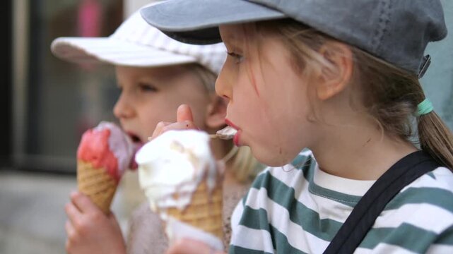 Two young girls savor ice cream cones outdoors, tasting melting dessert together. Sisters spend quality time side by side, showing warmth of sibling bonding. Each girl licks her cone.
