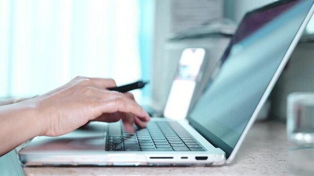 Close up of a hand tapping a smartphone screen and typing rapidly on a laptop keyboard at a home workspace, remote work and digital multitasking in a modern bedroom, condo or apartment interior.