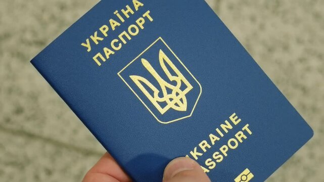 Ukrainian biometric passport in male hand near transport terminal floor. Blue travel document with gold national emblem and Ukraine lettering. International identification for border control and