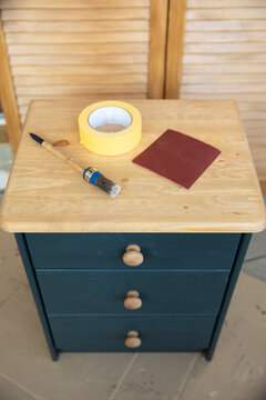 Top view of a small wooden cabinet undergoing restoration, with freshly painted dark blue drawers, a natural wooden top and painting materials arranged on the surface. The image illustrates a practica