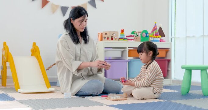Asian single mother claps gently while preschool daughter practices hands-on learning with colorful toy on mat, encouraging confidence, motor skill, patience, positive bonding, development at home