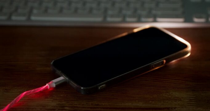 Close-up of a smartphone connected to a glowing charging cable on a wooden desk. 4K.