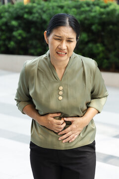 50s Southeast Asian woman feeling sick with stomach pain, grimacing and clutching her abdomen