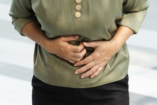 50s Southeast Asian woman feeling sick with stomach pain, grimacing and clutching her abdomen, hand closeup shot