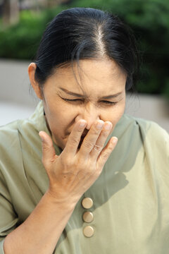 50s Southeast Asian woman gagging and nauseous in a park, covering her mouth with grimace expression