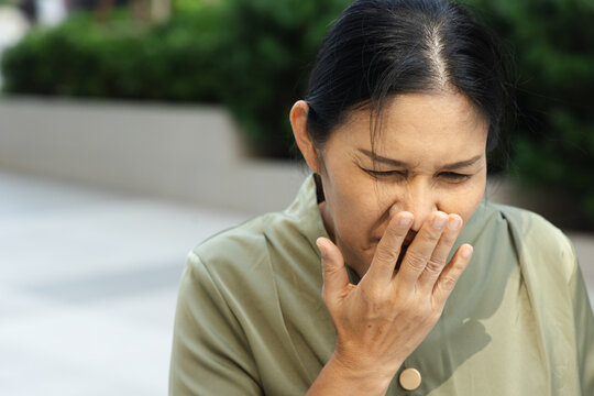 50s Southeast Asian woman gagging and nauseous in a park, covering her mouth with grimace expression