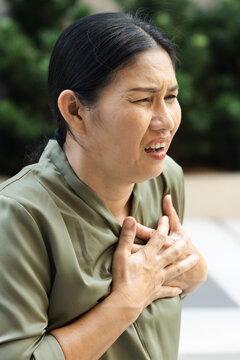Middle aged 50s Southeast Asian woman experiencing heart pain outdoors, hand on left chest and distressed expression