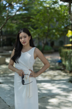 Asian woman in white dress holding sunglasses enjoying summer in green park