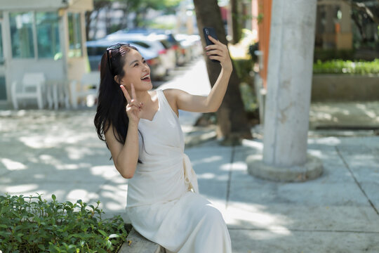 Asian woman taking outdoor selfie, smiling with v-sign gesture and enjoying sunshine