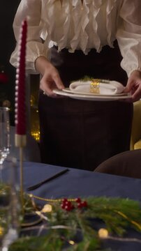 Vertical shot of woman carrying festive plates with diverse friends preparing Christmas dinner table. Suitable for holiday advertising campaigns
