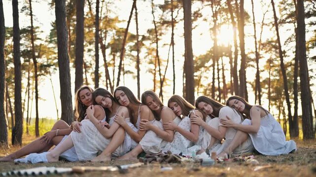 Women in white dresses sit closely together in a circle, embracing one another in a tranquil forest, with sunlight streaming through the trees