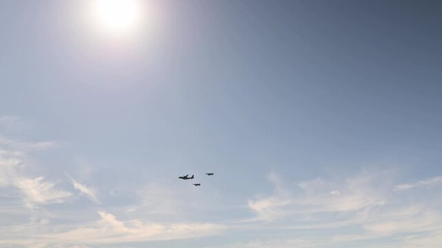 Two military fighter jets escort a large passenger aircraft flying in formation across a bright blue sky with thin clouds during a coordinated aviation flight in daylight overhead