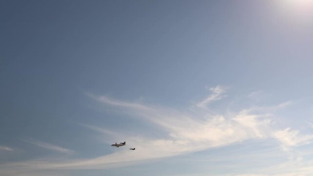 Two military fighter jets escort a large passenger airplane flying in formation across the bright daytime sky with soft clouds highlighting aviation security and air defense
