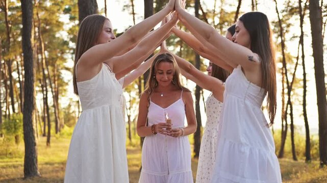 Group of young women in white dresses forming a circle around a central figure holding a candle in a forest setting during a sunny day