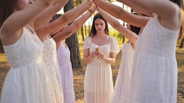 Young women in white dresses create a circle around a woman holding a candle in a forest, with sunlight filtering through the trees