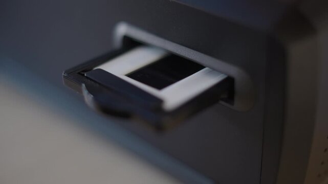 Close-up of a slide holder advancing through a film scanner as the scan starts, the scanner light faintly glowing through the housing. Digitising analog slides, a detail of film archiving at home, Ger