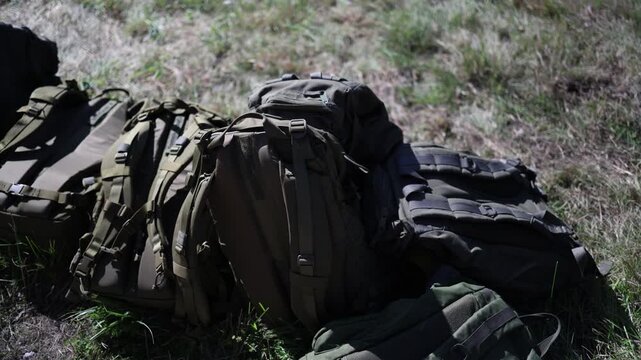 Military backpacks and tactical gear stacked on grass outdoors ready for deployment with rugged field packs and heavy duty equipment prepared for soldiers during field operations
