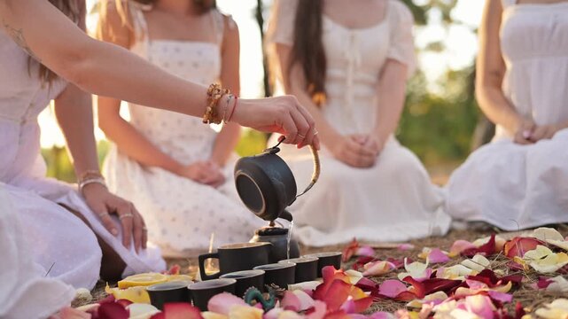 Women dressed in white engage in a group meditation, pouring tea from a kettle onto a tray surrounded by colorful rose petals in a natural environment