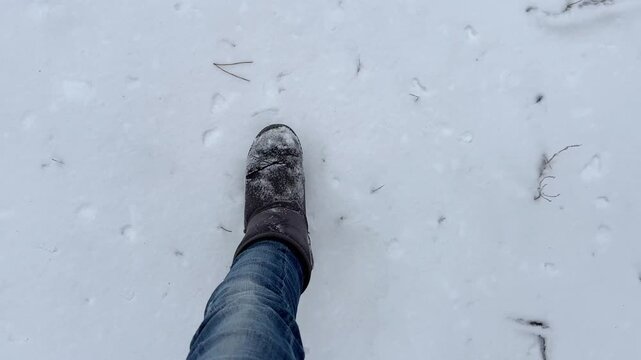 POV to female foot stepping on snowy path in park at cold day. Legs of young girl in uggs hiking through trail with white snow going at forest. Concept of winter active recreation and holiday trip.