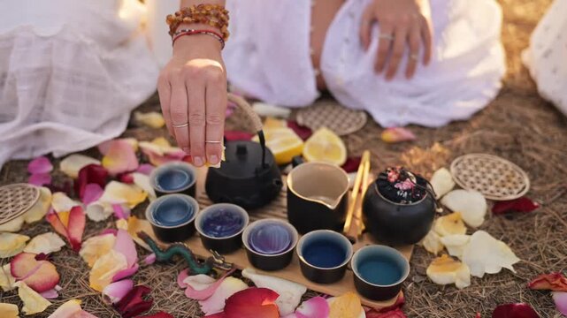 Woman pours tea from a black teapot into small cups placed among vibrant rose petals on a natural surface, with decorative elements in the background