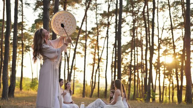 Woman in a flowing robe stands with a drum in a forest setting, surrounded by other women in white dresses engaged in meditation