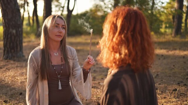 Two women converse in a sunlit forest, one wearing a dress and the other with curly red hair, surrounded by trees and natural scenery