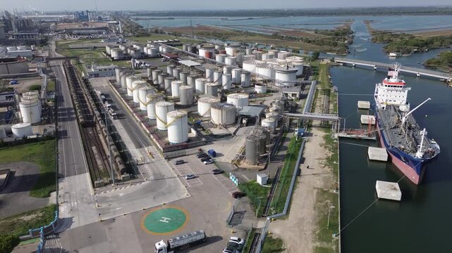 Smooth 4K drone flight over a port fuel terminal. Tanker ship moors at dock unloading into tanks. Railway with tank cars, trucks and helipad visible. High quality original unedited footage.