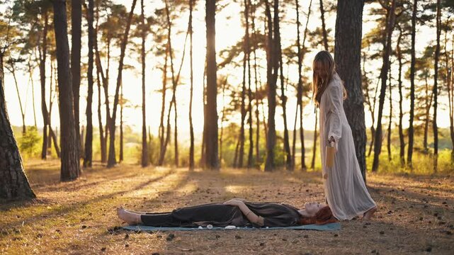 Woman in flowing robes conducts energy healing on another woman lying on a mat in a sunlit forest clearing with tall trees and natural surroundings
