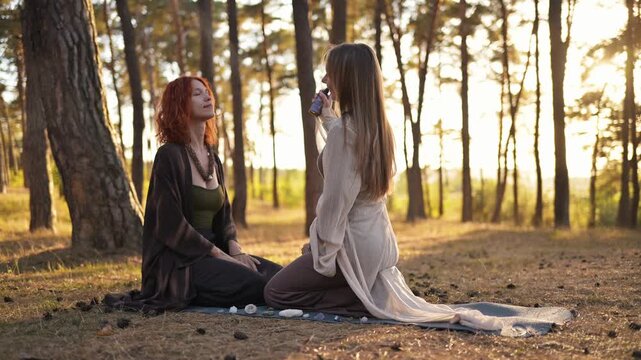Two women sit on the ground in a forest, one sprays mist on the other, both wearing flowing clothing, with trees and sunlight creating a serene atmosphere