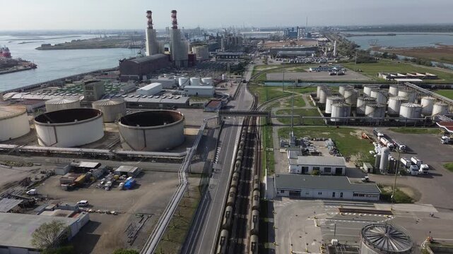 4K drone footage gliding over a port fuel depot with large storage tanks, rail line with tank cars, trucks and pipelines. Fuel plant with striped chimneys visible. Original unedited video.