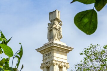 Cádiz's Constitution of 1812 Monument, featuring its central sculpture group and CADIZ inscription, standing under a clear blue sky, framed by vibrant green foliage © photoopus