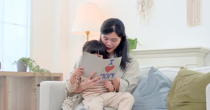Asian single mother holds preschool daughter on sofa while reading picture book at home, creating warm learning moment with language practice, emotional support, bonding, care, child development