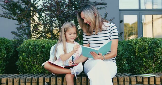 Mother and daughter sitting on bench sharing tablet and smiling in suburban garden during warm afternoon interaction, close eye-level family bonding scene on green hedge background. Back to school