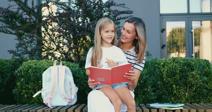Woman and daughter sharing storytime on bench in backyard with backpack and notebooks nearby, warm golden hour family moment of reading and laughter together. Back to school. Education concept.