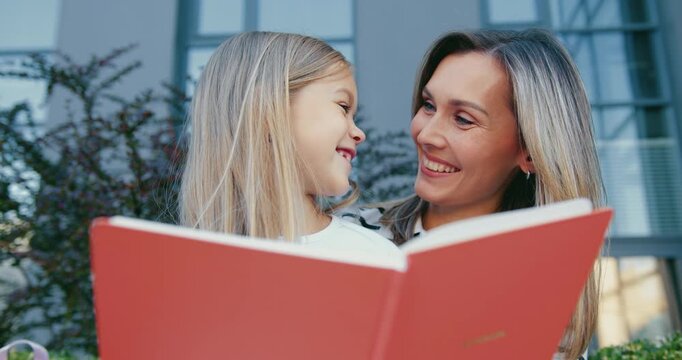 Mother and daughter reading large red storybook together on balcony at dusk, smiling and sharing warm family bond, close waist-up view with soft evening light. Back to school. Education concept.