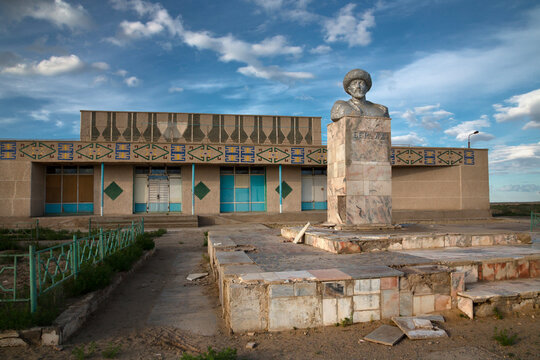 Stone bust of a bearded man in a fur hat on a marble pedestal in front of a decorated building with tiled patterns at dusk, Moynaq, Uzbekistan