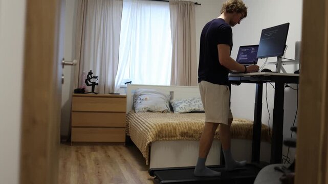 Young IT specialist walks on a treadmill while studying new technical diagrams and software systems on his computer combining professional education with daily exercise at home