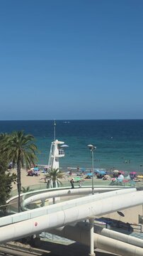 Alicante, Spain. - 30 August 2026 -  Elevated view of beachgoers relaxing on a sandy Mediterranean beach beside clear turquoise water on a sunny summer day in Alicante, Spain.