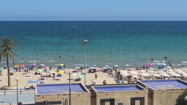 Alicante, Spain. - 30 August 2026 -  Elevated view of beachgoers relaxing on a sandy Mediterranean beach beside clear turquoise water on a sunny summer day in Alicante, Spain.