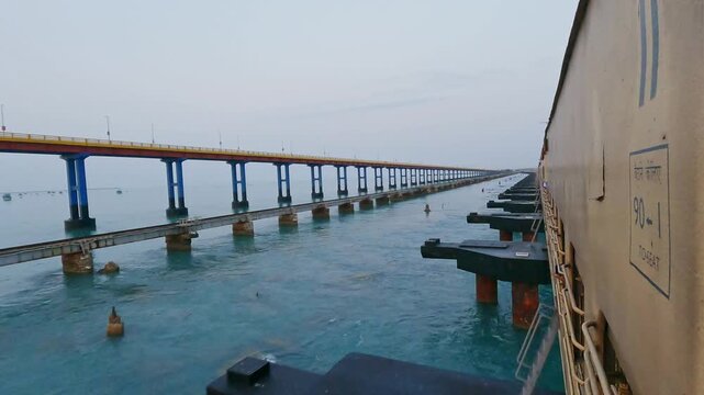POV video from a moving train crossing Pamban Railway Bridge near Rameswaram, Tamil Nadu, India, showcasing the railway tracks, turquoise seawater, and parallel bridge structure along the coast.