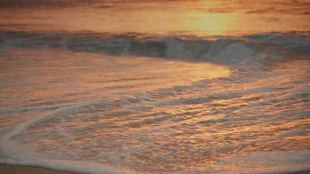 Ocean waves gently washing ashore on sandy beach during golden hour sunset creating relaxing warm seascape with soft foam and sun reflection