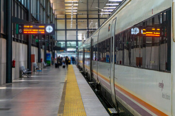 Train standing at a modern railway station platform in Cadiz, Spain, featuring a departure board reflection on its windows and travelers walking with luggage, symbolizing travel, transportation.  © photoopus