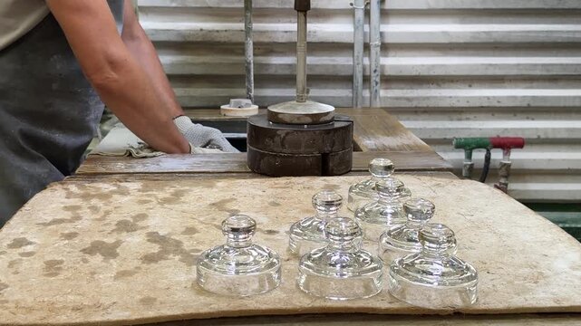 Clear glass lids with rounded knobs rest on a worn workbench beside a gloved artisan in a glassmaking workshop