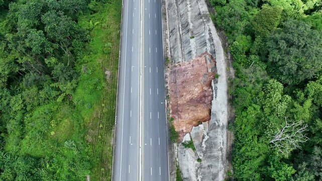 Aerial view captures a major landslide damaging a concrete roadside slope, revealing exposed red soil, structural failure, erosion risks, and transportation safety concerns. Nan Province, Thailand.
