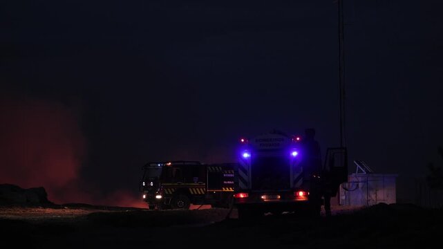 Firefighters and vehicles illuminated by flashing lights respond at night to a wildfire, with thick smoke and orange haze filling the rural landscape during a summer heatwave crisis.