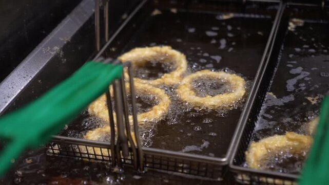 Golden battered onion rings sizzle in deep fryers as bubbling oil crisps the coating in a bustling restaurant kitchen. Close-up view captures texture, movement, and rich color.