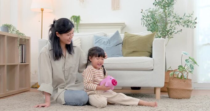 Asian single mother watches preschool daughter hold pink piggy bank after saving practice on carpet at home, encouraging money learning, independence, confidence, nurturing family bond through play.